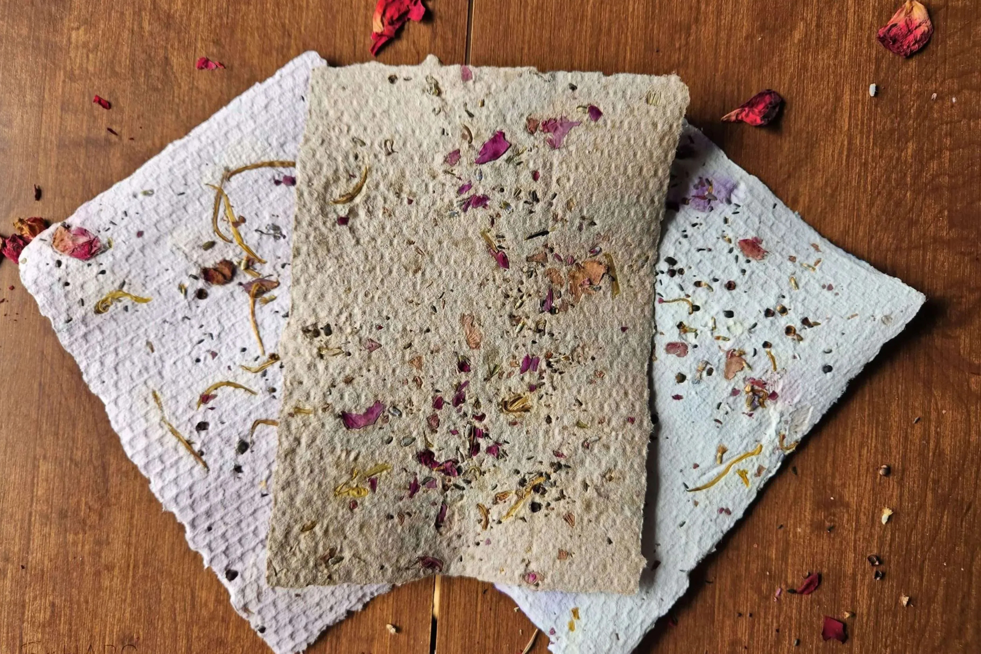 The photograph shows three sheets of handmade paper embedded with seeds and dried purple flowers. They are placed on a wooden surface and surrounded by purple-red flowers.