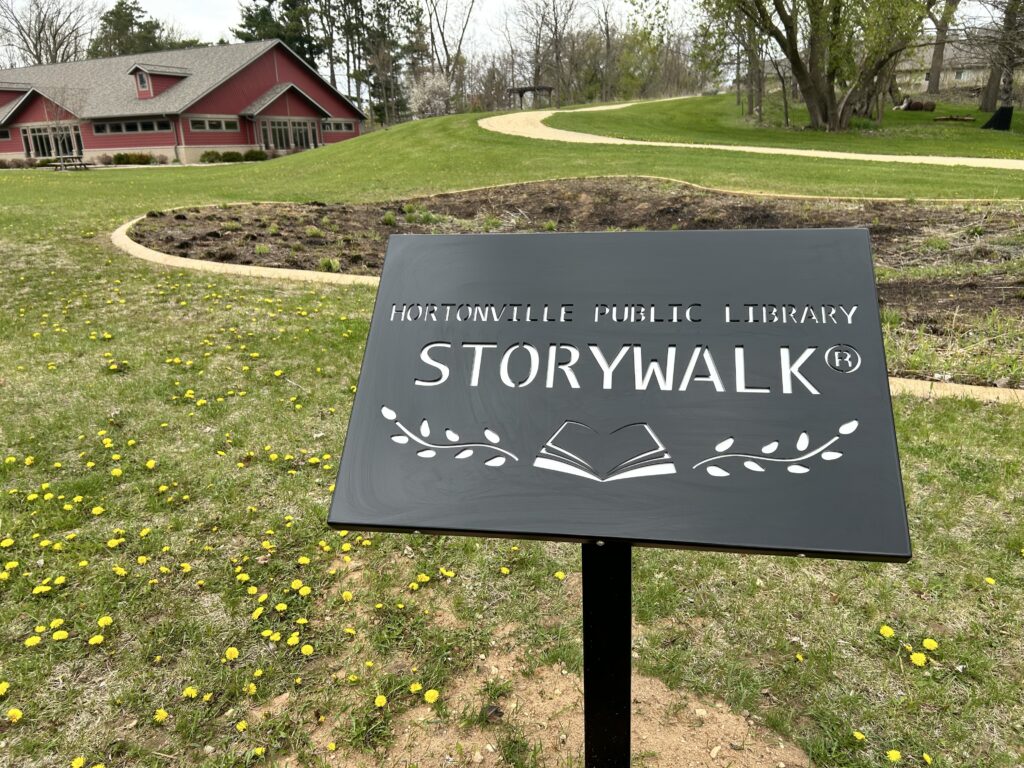 Black metal sign says "Hortonville Public Library StoryWalk" with a background of a gravel walking path and the outside of the Hortonville Public Library.