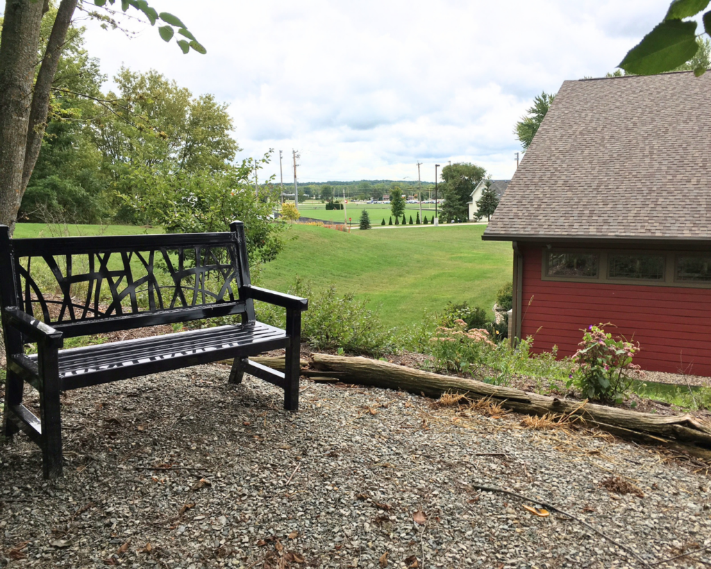 A metal bench on a gravel trail overlooks the back of a red building- the Hortonville Public Library.